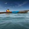 A young child sitting in a small boat on a bright blue sea, surrounded by sparkling waves under a clear, sunny sky. The child is wearing a colorful life jacket as well